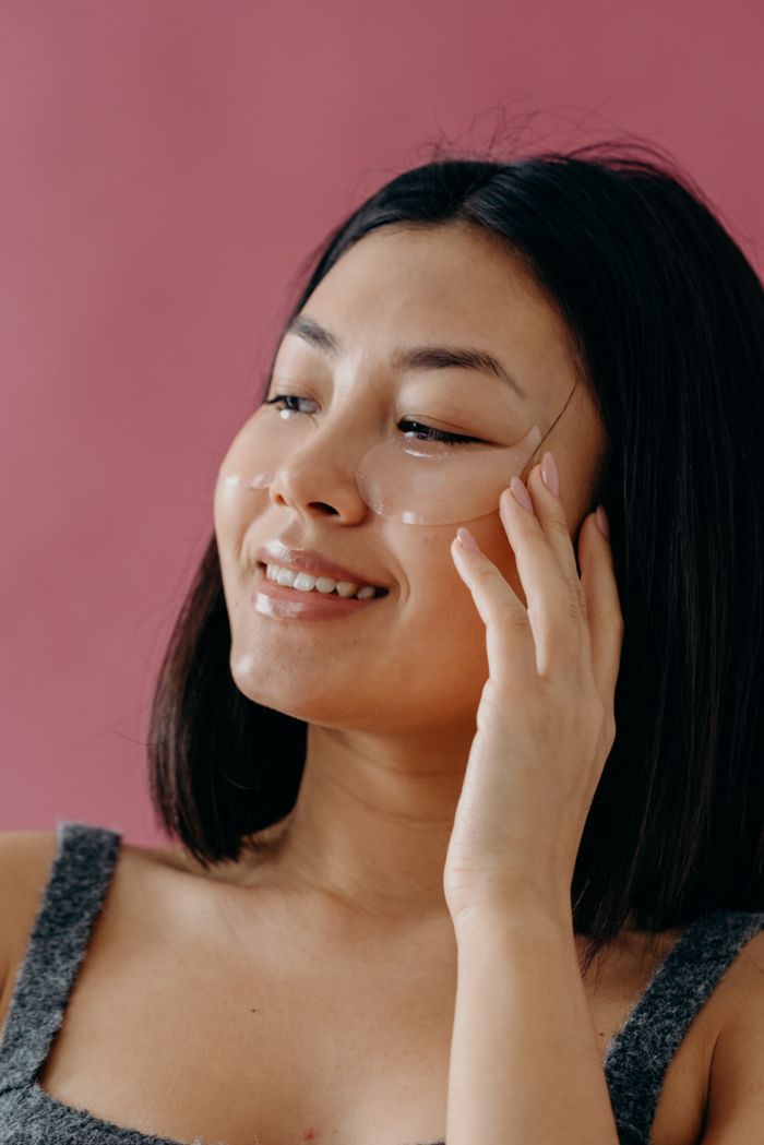 Smiling Asian woman applying under eye patches for skin care against a pink background.